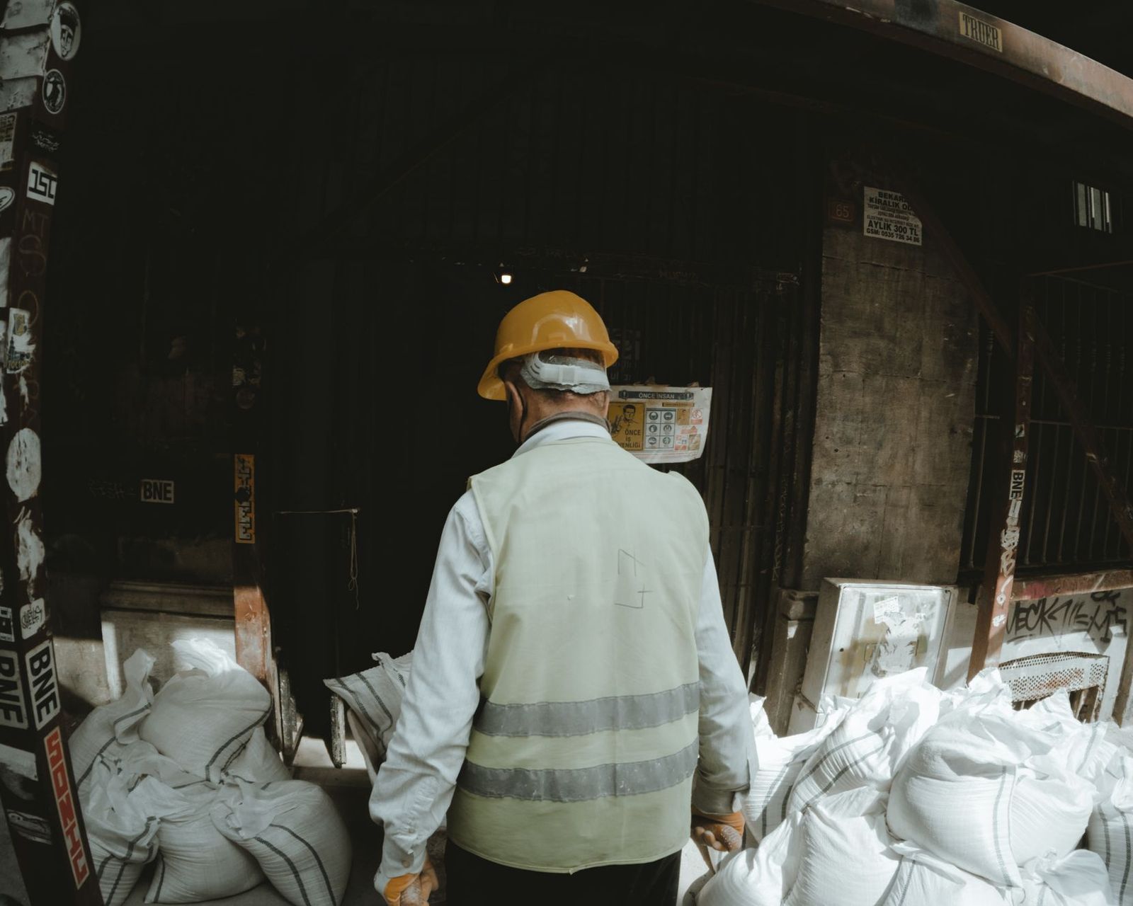 Trabalhador de costas, com capacete e colete refletivo, dentro de um galpão